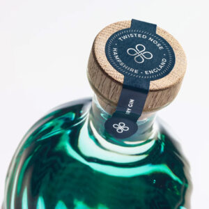 Twisted Nose Gin è un gin secco pluripremiato prodotto nell’Hampshire, pensato per racchiudere la purezza e la freschezza dei corsi d’acqua calcarei (chalk streams) che scorrono vicino alla distilleria. Realizzato in piccoli lotti e distillato lentamente in alambicchi di rame, nasce per valorizzare la provenienza e il legame con un habitat naturale unico. La firma aromatica è il crescione fresco locale, naturalmente pepato, che dà al distillato un tocco stimolante e un finale vivace e persistente; il riferimento storico richiama il nome romano “Nasturtium”, interpretato come “naso storto”. Accanto al ginepro, una selezione di botaniche completa il profilo con equilibrio: pompelmo, corteccia di cassia, semi di finocchio, iris, radice di angelica, lavanda, agrumi e coriandolo, distillate con cura per mantenerne gli aromi freschi e delicati. Al naso risulta morbido, con sfumature erbacee e un accenno speziato; al palato è fresco, secco e ben bilanciato, con note di pompelmo e finocchio, cenni delicatamente floreali e una chiusura pepata distintiva legata al crescione. La persistenza è lunga e pulita, con una sensazione complessiva intensa ma mai pesante. Anche il design è parte dell’esperienza: la bottiglia personalizzata in vetro blu-verde presenta un effetto ondulato che increspa e rifrange la luce come la superficie di un ruscello, anticipando visivamente la sua anima “di fiume”. In servizio è ideale in Gin Tonic con una fetta di pompelmo rosa per esaltare la componente agrumata, ed è eccellente anche in Martini con vermouth, dove la sua secchezza e la traccia erbacea emergono con grande precisione.