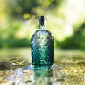 Twisted Nose Gin è un gin secco pluripremiato prodotto nell’Hampshire, pensato per racchiudere la purezza e la freschezza dei corsi d’acqua calcarei (chalk streams) che scorrono vicino alla distilleria. Realizzato in piccoli lotti e distillato lentamente in alambicchi di rame, nasce per valorizzare la provenienza e il legame con un habitat naturale unico. La firma aromatica è il crescione fresco locale, naturalmente pepato, che dà al distillato un tocco stimolante e un finale vivace e persistente; il riferimento storico richiama il nome romano “Nasturtium”, interpretato come “naso storto”. Accanto al ginepro, una selezione di botaniche completa il profilo con equilibrio: pompelmo, corteccia di cassia, semi di finocchio, iris, radice di angelica, lavanda, agrumi e coriandolo, distillate con cura per mantenerne gli aromi freschi e delicati. Al naso risulta morbido, con sfumature erbacee e un accenno speziato; al palato è fresco, secco e ben bilanciato, con note di pompelmo e finocchio, cenni delicatamente floreali e una chiusura pepata distintiva legata al crescione. La persistenza è lunga e pulita, con una sensazione complessiva intensa ma mai pesante. Anche il design è parte dell’esperienza: la bottiglia personalizzata in vetro blu-verde presenta un effetto ondulato che increspa e rifrange la luce come la superficie di un ruscello, anticipando visivamente la sua anima “di fiume”. In servizio è ideale in Gin Tonic con una fetta di pompelmo rosa per esaltare la componente agrumata, ed è eccellente anche in Martini con vermouth, dove la sua secchezza e la traccia erbacea emergono con grande precisione.